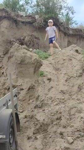 A young child wearing a light-colored t-shirt, dark shorts, and a baseball cap is actively climbing a steep, sandy embankment. The child is on all fours, moving upwards, with a determined expression. The embankment appears to be made of loose soil and rocks, with some sparse green vegetation growing on its slopes and at the top. Behind the child, the embankment continues upwards, with a denser growth of trees and bushes forming a natural barrier against a cloudy sky. The overall setting is outdoors, with a natural, undeveloped landscape. In the foreground, the edge of a trailer is visible, with a dark tire and part of its metal frame, suggesting that the child might have been dropped off or is exploring near a vehicle. The lighting suggests it is daytime, possibly late morning or afternoon. The child's posture and facial expression convey a sense of focused effort and adventurous play. The location is identified as Bel'tirskiy, Russia.