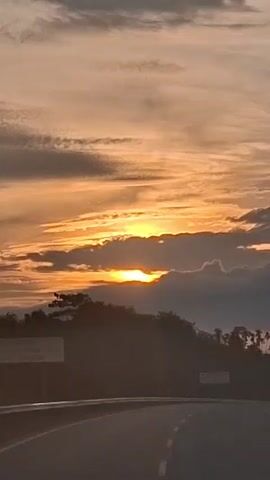 The video shows a view from a moving vehicle at sunset. The sun is partially obscured by clouds and is casting an orange glow across the sky. There are trees and hills in the foreground, with a road curving in front of the vehicle. There are also some signs visible by the side of the road. The overall mood is serene and peaceful.