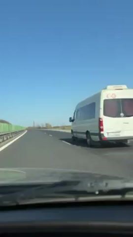 The video shows a car driving on a highway on a clear, sunny day. A white van is briefly seen driving in the right lane before it speeds up and out of view. The highway has two lanes and is bordered by a green fence on the left and a grassy field on the right. The sky is bright blue. The perspective is from inside the car looking out the windshield.