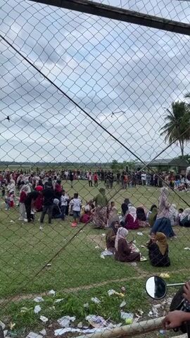 The video shows a large gathering of people outdoors, viewed through a wire mesh fence. A crowd of people, some seated and some standing, are spread across a grassy area. Many are wearing headscarves and traditional clothing. In the background, there are more people, some tents, and palm trees under a cloudy sky. The foreground shows the wire fence and some debris on the ground, including plastic and paper waste. A motorcycle mirror is visible in the bottom right corner. The overall impression is of an outdoor event or gathering with a substantial number of attendees.