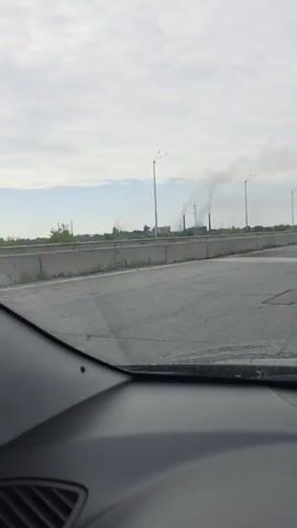 The video shows a view from inside a vehicle traveling on a highway. The road is visible in the foreground, with a concrete barrier on the right. In the distance, there is an industrial complex with several smokestacks emitting smoke. A black Volkswagen Polo hatchback is seen driving on the highway to the right of the vehicle from which the video is being recorded. The sky is overcast.