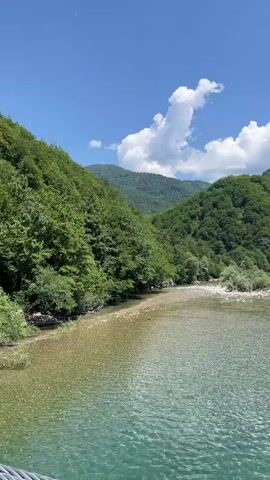 The video captures a scenic view of a wide, clear river flowing through a lush green valley. The water is a beautiful turquoise, with visible pebbles and aquatic plants beneath the surface in the shallower areas. The riverbanks are lined with dense, vibrant green trees and vegetation. In the background, rolling hills covered in more trees rise up, creating a majestic mountainous landscape. The sky is a bright blue, dotted with fluffy white clouds, indicating a clear, sunny day. The camera pans slowly across the scene, offering a panoramic perspective of the natural beauty. There are no people or man-made objects immediately visible in the river or on the immediate banks, suggesting a remote and natural setting. The overall impression is one of tranquility and natural splendor.