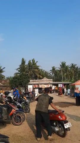 A man in a dark vest and black pants pushes a red scooter forward. He is facing away from the camera. Several other scooters are parked in a row to his left. People are visible in the background, walking and standing near a row of market stalls. Palm trees and houses are visible behind the stalls. The sky is clear and blue.