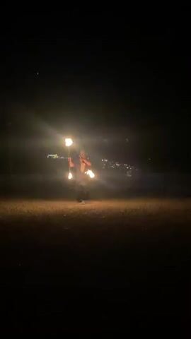 The video shows a person performing with fire in a dark, outdoor environment. In the first clip, a person is visible in the distance, holding what appears to be a torch with flames. The second clip shows a close-up of a large fire, possibly from a flamethrower or a large torch, with the person operating it mostly obscured by the flames. The third, fourth, and fifth clips return to the person from the first clip, who is now closer and appears to be juggling or manipulating multiple flaming objects, creating light trails in the darkness. In the background, a faint, distant town or city with lights is visible. The ground is covered in what looks like dry grass or dirt. The overall scene suggests an evening or night performance.