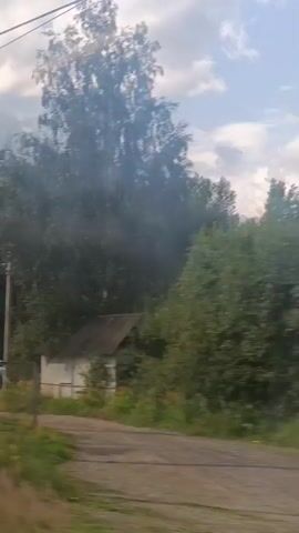 The video shows a view from a moving vehicle, looking out at a rural landscape with trees and a railway crossing. In the foreground, there is a metal gate and some grass. Beyond that, a white SUV is visible near a railway track. A railway crossing gate is down, indicating that a train may be approaching or passing. The sky is cloudy, and the overall impression is of a bright, sunny day.