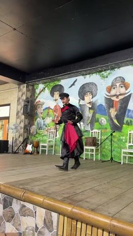 A man in traditional attire is dancing on a wooden stage in front of a mural. The mural depicts sheep, stylized figures, and a landscape. The man is wearing a black coat with a red sash and black pants tucked into boots, and a black hat. He is moving his arms and legs in a dynamic way. White chairs and drums are placed on the stage behind him. There is a speaker and a fan to the left of the stage.