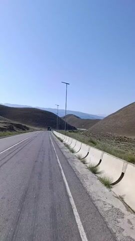 The video shows a view from a vehicle traveling on a paved road with a white lane marking. The road curves to the left, with gently sloping, dry, brown hills on either side. There are sparse patches of green vegetation on the hillsides and along the side of the road. A concrete barrier with arched openings runs along the right side of the road. Several tall, modern streetlights with solar panels are spaced along the road. In the distance, a truck can be seen traveling in the same direction. The sky is clear and blue. The overall environment appears to be a rural, arid landscape on a sunny day.