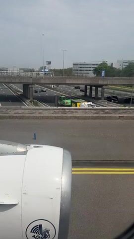 The video shows the view from an airplane window on the ground. The wing and engine of the plane are visible in the foreground. Beyond the runway, a highway runs parallel with the runway, with cars traveling in both directions. There is an overpass bridge crossing the highway. In the background, there are buildings and signage visible. The weather appears to be overcast.