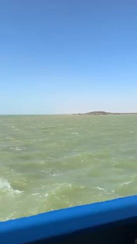 The video features a view from the deck of a boat traversing a large body of water on a sunny, clear day. The sky is a bright blue, meeting a horizon where low-lying, arid-looking land is faintly visible in the distance. The water itself is a distinct greenish-brown color, with noticeable waves indicating movement and potentially wind.

The perspective shifts between a direct view over the water, showing a blue railing in the foreground, and wider shots of the boat's deck. These wider views reveal the boat's structure, including white support pillars, an overhead mesh canopy, and various industrial elements. On the deck, blue-painted pipes, black cylindrical bollards with yellow tops, and coiled ropes are visible. A person wearing a red outfit can be seen partially on the deck, standing near some equipment, suggesting a crew member. The boat is in motion, cutting through the wavy water. No text is visible in the video.