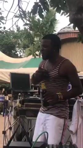 A man with dark skin, facial hair, and wearing a striped tank top and light-colored pants, is singing into a microphone and holding a bottle of beer. He is standing in front of a DJ setup with a laptop and mixer. The background includes tents, trees, and people sitting at tables. The environment appears to be an outdoor event.
