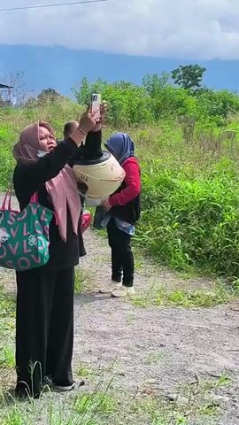 The video shows a wide shot of a steep, eroded landscape. In the foreground, a person wearing a hijab, red long-sleeved shirt, and dark pants is walking away from the camera, carrying a light-colored bag. To the right, a portion of a dark-colored vehicle is visible. The landscape itself features dramatic gullies and cliffs with sparse vegetation. In the background, hills and trees are visible under a cloudy sky. The erosion appears significant, creating a rugged and uneven terrain.