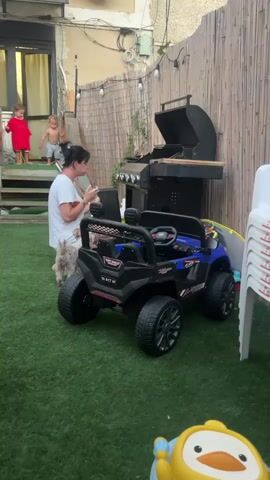 A woman kneels on artificial grass in a backyard, facing a toy jeep and a grill. Two young children, one wearing a red shirt and the other shirtless, are visible on a set of stairs in the background. The woman is holding something in her hands. The backyard is fenced with a bamboo-style screen and string lights are visible overhead.