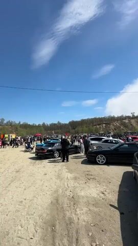 The video shows a crowd of people at an outdoor car show on a sunny day. The sky is blue with scattered clouds. Numerous cars, mostly vintage Japanese models like Toyota Supras and Nissan Skylines, are parked on a large dirt lot. People are walking around, looking at the cars and talking. A few small tents and other temporary structures are visible in the background. The surrounding terrain includes low, tree-covered hills.