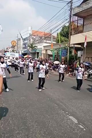 The video shows a group of young women in matching white and blue athletic tops with black pants and black headscarves performing a choreographed dance routine on a paved street. They are moving in formation, with some individuals in the foreground and others further back in a line. The street is lined with buildings and there are spectators on the sidewalks. There is a white van with a banner on it in the background. The sky is bright and clear, suggesting it is daytime. The overall scene appears to be a public event or parade.