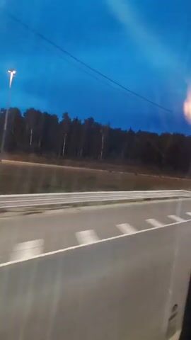 The video shows a view from inside a vehicle, likely a bus or car, looking out through a window at dusk or night. The sky is a deep blue with some lighter hues near the horizon. A line of trees forms a dark silhouette against the sky. A street or road is visible in the foreground, with white lane markings illuminated by the vehicle's headlights and possibly streetlights. The vehicle appears to be moving, as indicated by the motion blur. The camera briefly pans to the side, showing the interior of the vehicle, with reflections on the window. The overall scene suggests travel at night on a road passing through a wooded area.