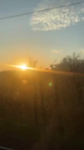 This video shows a sunset viewed through a window. The sun is low on the horizon, casting a warm golden light across the sky. Clouds are visible, tinged with orange and pink. The foreground is blurred, suggesting movement, perhaps from a vehicle. Trees and other vegetation are also visible, appearing as dark silhouettes against the bright sky. The overall impression is one of a peaceful and serene moment, perhaps during a journey.