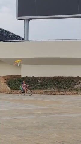 A video shows a woman and a child riding a bicycle and flying a kite on a large, open, paved area. The woman is wearing a pink top and a red cap, while the child, sitting behind her, is in a green shirt and a blue hat. They are riding a white bicycle, and a yellow kite with colorful streamers is flying high in the sky above them. The backdrop features a modern building with a large electronic display screen and a small green embankment. The sky is overcast, and the environment looks spacious and relatively empty.