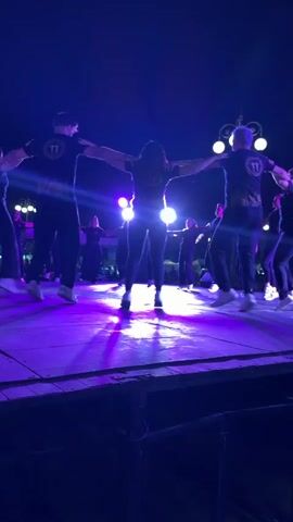 A group of about ten people are line dancing on a stage. They are all wearing dark clothing, and the men appear to be wearing shirts with a logo on the back. The stage is lit with purple lights and there are light stands in the background. The dancers are holding hands or arms in a line, and they are all facing away from the camera. The sky is dark.