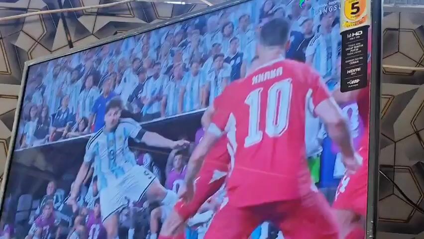 The video shows a television screen displaying a soccer match. The screen is framed by a patterned wallpaper. The match appears to be a close-up of players in red jerseys facing players in white jerseys. The crowd in the background is wearing white. The ball is in the air, and a goalkeeper in a black and white striped jersey is jumping to catch it. The text "WORLD CUP" is visible on a banner in the background. There is also an electronic banner on the right side of the screen with text and logos, including "UHD" and "HDMI". The lower part of the television screen has the brand name "POLYTRON".
