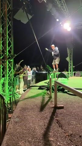 A man is standing on a stage at night, holding a drink in his left hand and a microphone in his right. He is wearing a baseball cap, a dark jacket, and plaid shorts. He is facing a crowd of people who are standing behind a metal barrier. The stage is lit with green and white lights. There are speakers and lighting equipment on and around the stage. The crowd is visible in the background, with some people holding up their hands. The man appears to be performing or speaking to the crowd.