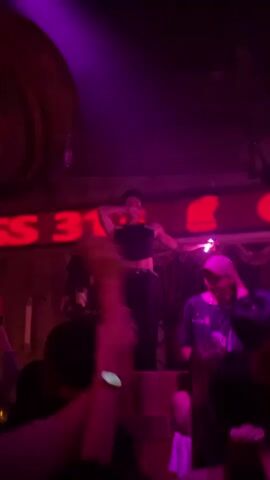 A person is performing on a stage in a dimly lit room. The performer is wearing a black crop top and black pants and is holding a pink glow stick. There is a neon sign behind the performer that says "ASS 313". The room appears to be a club or bar, with a crowd of people watching the performance. The lighting is predominantly red and pink.