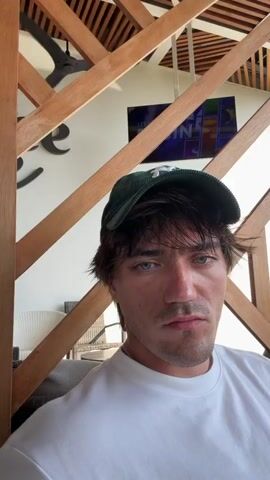 A man in a green cap and white t-shirt sips from a wine glass. He winks at the camera. The man is sitting on a patio of sorts with wood lattice and a sign reading "Golf Zone & Games". He is looking out at a golf course. Another man in shorts is practicing his golf swing.