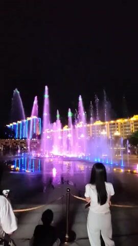 The video shows a night scene of a water fountain show. The fountain is illuminated with colorful lights, primarily purple and blue, with some green highlights. The water jets are shooting upwards and creating various shapes. In the foreground, there are three people. Two are in the middle ground, a child sitting down and a person standing, both facing the fountain. In the left foreground, a person is partially visible, holding a phone and facing the fountain. In the background, there are buildings with glowing lights, suggesting an urban environment. The sky is dark, indicating it is nighttime. The fountain appears to be synchronized with music or a light show.