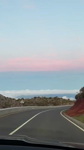 The video shows a car driving on a mountain road towards the horizon. The road is paved with black asphalt and has a single solid white line dividing the lanes. On the left side of the road is a low concrete barrier, behind which is a landscape of dense, low-lying vegetation. On the right, the roadside slopes up with reddish-brown soil and some sparse greenery. Above this vegetation is a telephone pole with wires running across the scene. The sky is a gradient, transitioning from a light blue at the top to a band of pinkish clouds nearer the horizon, beneath which is a layer of dense white clouds resembling a sea of fog. The camera's view is from inside the car, with the dashboard visible at the bottom of the frame.