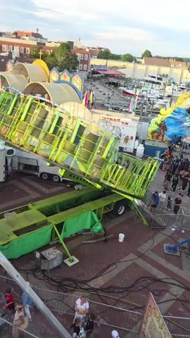 This video shows an aerial view of a carnival ride, likely a spinning or tilting amusement ride, in a setting with buildings and boats visible in the background. The ride is primarily green and yellow, with lights. It appears to be in operation or being set up, as a section of it is elevated. People are visible on the ground near the ride and in the background. The sky is cloudy.