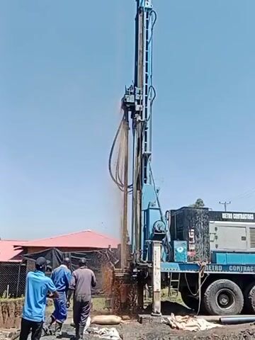 The video depicts a water drilling operation. A large, blue drilling rig, labeled "Metro Contractors Drilling," is positioned over a muddy area, actively drilling into the ground. Water and mud are being ejected from the drill site. Three people are present, presumably workers. One is standing near the rig, observing the drilling. The environment appears to be an outdoor area, possibly a construction site or undeveloped land, with a red-roofed structure visible in the background and a clear, blue sky overhead.