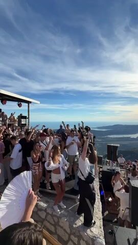 This video shows a large group of people dancing and enjoying themselves at an outdoor party. The party appears to be held on a mountaintop or high elevation, with a stunning panoramic view of a bay and surrounding mountains. The sky is blue with scattered clouds, and the sun is visible, casting a warm glow. The crowd consists of men and women dressed in casual, summery attire, many of them raising their hands in the air and dancing to music. There are speakers set up, indicating a DJ or music system is in place. Some individuals are holding drinks, and a white fan is visible in the foreground, held by one of the attendees. The overall atmosphere is festive and energetic, set against a breathtaking natural landscape.