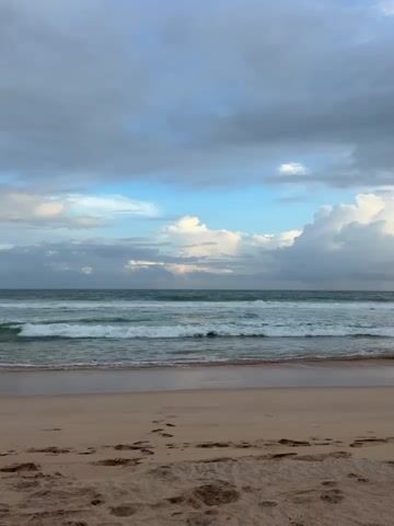 The video captures a serene beach scene with the ocean in the background and sand in the foreground. The sky is a mix of blues and grays with scattered clouds. The ocean has gentle waves rolling towards the shore, and the water appears to be a greenish-blue color. The sand is light brown and textured, with several footprints visible. The overall impression is one of a calm and natural environment, likely during daylight hours, though the lighting suggests it could be early morning or late afternoon. There are no people or objects clearly visible in the frame, focusing solely on the landscape.