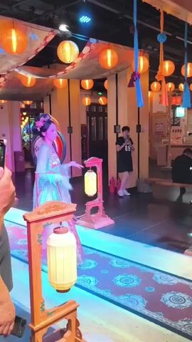 A woman in traditional Chinese attire, adorned with a floral headdress, performs a dance on a stage illuminated with blue and purple lights. She is dressed in a flowing blue and pink gown with long sleeves and intricate embroidery. The stage is decorated with red and orange lanterns suspended from the ceiling, creating a festive atmosphere. In the foreground, two people are visible from behind, watching the performance. The person on the left is holding a phone, capturing the dance. In the background, other individuals can be seen sitting or standing, also observing the performance. The overall setting appears to be an indoor venue decorated in a traditional Chinese style, possibly a restaurant or entertainment space.