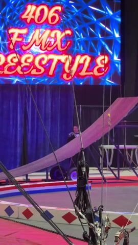 The video captures an indoor stunt show performance. The initial shot shows a stage with a large LED screen displaying "406 FMX Freestyle" in red and blue letters against a dynamic blue background. Below the screen, a metal ramp angled upwards is visible, with a person standing near its base. Numerous ropes and cables hang down in the foreground.

As the video progresses, a quad bike (ATV) is seen launching from a ramp and flying through the air. The quad bike is white and blue, and the rider is not clearly visible during the aerial maneuver. The background behind the flying quad bike features several advertising banners, including "TRICART Ford", "SWEETCORN FESTIVAL", "AUDIO Visual", and "Maddin Motorcycle Club".

The quad bike descends and lands on a large, black, inflatable landing pad. The landing pad also features the "406 FMX Freestyle" logo in white. In the foreground, a man wearing a blue jacket and a red fez is partially visible, observing the stunt. The entire event appears to be taking place in a large arena or exhibition hall, indicated by the high ceiling and visible truss structures with lighting. The atmosphere seems to be exciting and energetic, characteristic of a live performance.