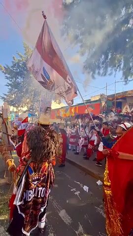 The video shows a parade from a low angle, focusing on a person in a vibrant costume and a large flag. The person in costume is wearing a red jacket, a white and red skirt-like garment with intricate designs, white leggings with red accents, and black boots. They have a headdress with dark, bushy hair and wear a straw hat. The flag, held by a pole, is predominantly white with red accents and features a graphic of a bird or creature and text that appears to read "PABITOS". A red umbrella-like object is visible on the right side of the frame. In the background, there's a wooden float or structure adorned with greenery, possibly part of a procession, and other people are visible, some in similar festive attire. Smoke or colored powder, possibly red, is dispersed in the air, adding to the celebratory atmosphere. The environment appears to be an outdoor street with trees and buildings in the distance.