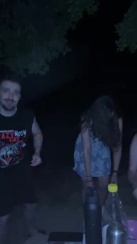 A man in a black sleeveless "CRAZY MEOW CAT" t-shirt and black shorts dances animatedly in a dimly lit outdoor environment at night. He has a beard, mustache, and red strings wrapped around his arms. He smiles widely, gestures with his hands, and briefly sticks out his tongue. Behind him, a woman with long, dark, curly hair, wearing a light-colored patterned dress, is bent over and moving. Hands of other individuals are visible in the bottom left foreground. On the bottom right, a dark water bottle, a clear bottle with a yellow cap, a metallic container, and a smartphone are visible on a surface.