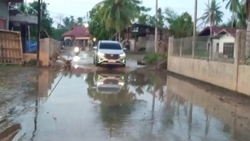 A silver SUV drives through a flooded rural road. The water is knee-deep and the car is kicking up a large splash as it navigates the submerged road. The sky is overcast and it appears to be dusk. There are houses and trees on either side of the road. The car's headlights are on, and its reflection is visible in the water.