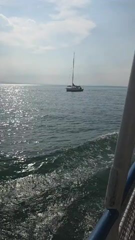 The video shows a view from a moving boat on the water. In the distance, there is a sailboat with a tall mast. The water is choppy, with waves and white foam trailing behind the boat. The sky is clear and blue with some clouds. The sun is shining, creating reflections on the water. There are other boats visible in the distance. The camera is angled slightly downwards, showing the side of the moving boat in the foreground.