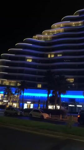 The video shows a nighttime view of two modern, wave-shaped buildings illuminated with blue and white lights. The buildings have many floors and balconies. Palm trees are visible in front of the buildings, and there are streetlights and cars on the street in the foreground. The overall impression is of a luxurious, contemporary urban environment at night.