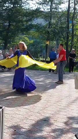 A woman in a blue dress and yellow fabric is dancing outdoors on a paved area. She is holding out large yellow pieces of fabric that extend from her arms, creating a wing-like effect. She is twirling and moving with the fabric. In the background, there are trees and several people sitting and standing, observing the dancer. The lighting suggests it is daytime.