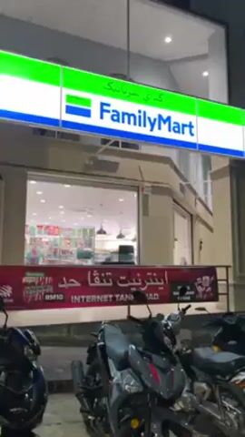 The video shows the exterior of a FamilyMart convenience store at night. The store's sign is prominently displayed, with the name "FamilyMart" written in English and another language above it. Below the sign, a banner advertises internet packages. Several motorbikes are parked in front of the store. The store's interior is visible through the large glass windows, with shelves stocked with various products. There are no people visible in the shot.