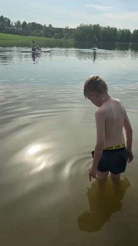 A young boy with short blonde hair and black swim trunks stands waist-deep in a lake. He turns to face the camera and smiles, winking. There are two people on paddle boards in the background, a green, grassy hillside, and a cloudy, blue sky. Sunlight reflects off the water.