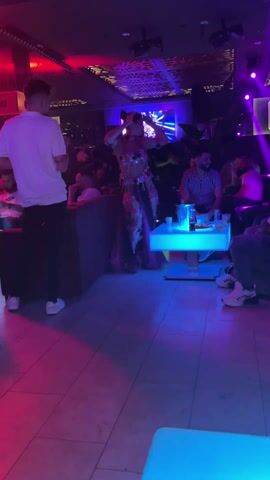 The video shows a woman in a sequined belly dancing outfit dancing in a crowded nightclub. She has her hands above her head and is swaying her hips. The lighting in the club is dark, with lots of pink and blue spotlights. Other people are sitting at tables around her, drinking and talking. There is a video screen visible in the background. Some people in the audience are giving her high-fives. The flooring appears to be tile.