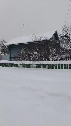 The video shows a snowy landscape in a village or rural area. There's a road or pathway covered in snow with tire tracks visible. On either side of the road, there are houses with snow-covered roofs. The houses vary in color, including a reddish-orange one and a gray one. The houses are surrounded by fences, some made of wood and some of metal. Trees and bushes, devoid of leaves in the winter, are scattered throughout the scene. The sky is overcast and gray, suggesting a cold, wintry day. At one point, a small dog briefly appears in the frame. The camera appears to be moving along the road, providing a view of the winter landscape.