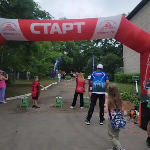 The video shows an outdoor race event. At the beginning of the video, a red inflatable archway with the word "СТАРТ" (START) printed on it is visible. Several people are standing around, some wearing red vests. A group of runners is in the distance, approaching the start/finish line. As the video progresses, a female runner wearing a purple top and shorts crosses the finish line. A man in a blue t-shirt and black pants, who appears to be an event official or volunteer, congratulates her. There is a tripod with a timer or display screen near the finish line, and some cardboard boxes and equipment are present. Trees and bushes form the background, and a building is visible on the right side of the frame. The overall atmosphere appears to be one of a community sports event.