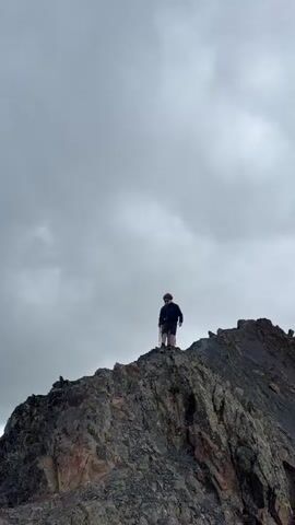 A video shows two people standing on a rocky mountain ridge under a cloudy sky. The person in the foreground is wearing a dark jacket and shorts and is holding trekking poles. The person in the background is also wearing dark clothing. The camera is positioned below the ridge, looking up at the individuals.