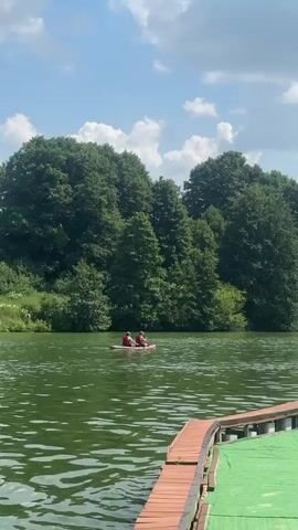 The video shows a lake surrounded by trees under a blue sky with white clouds. In the distance, two people are in a small red boat on the water. In the foreground, there is a wooden dock with a green surface. The camera pans to the right, showing a person lying on a lounge chair on the dock.