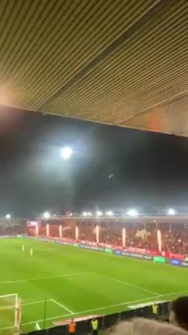 The video shows an outdoor soccer stadium lit up at night, with people filling the stands. Pyrotechnics are set off along the edge of the field, creating flames and smoke in front of the crowd. The camera is positioned from within the stadium, looking out over the field and up at the stadium's roof structure. The stadium is crowded, suggesting a significant event.