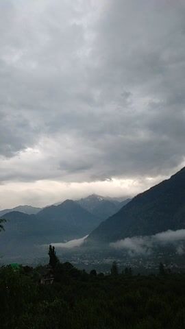 This is a vertical video of a mountainous landscape in Patar, India. The scene is dominated by lush green vegetation on the left, cascading down a steep slope. Below the vegetation, a valley with a village or town can be seen, partially obscured by mist or low-lying clouds. The mountains themselves are dark and imposing, with layers of peaks receding into the distance. The sky is overcast with heavy, grey clouds, suggesting a potentially rainy or stormy day. The light filtering through the clouds suggests it is either early morning or late afternoon. There are no people or distinct objects visible, only the natural landscape. The overall mood is serene and perhaps a little dramatic due to the weather. The image is visually striking due to the contrast between the vibrant green foliage and the muted tones of the mountains and sky. There is no visible text in the video.