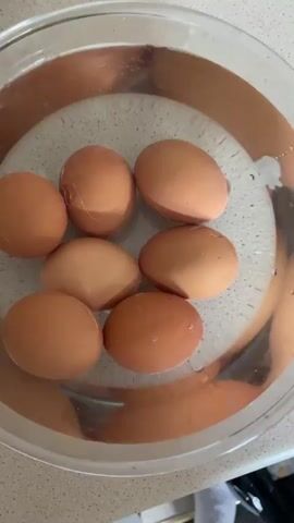 The video shows a top-down view of a clear glass bowl filled with water. Inside the bowl are seven brown eggs. The eggs are submerged in the water and arranged in a rough circular pattern. One egg in the upper left appears to have a small crack on its shell. The bowl is sitting on a light-colored speckled countertop. The lighting appears to be natural, coming from above and slightly to the right.